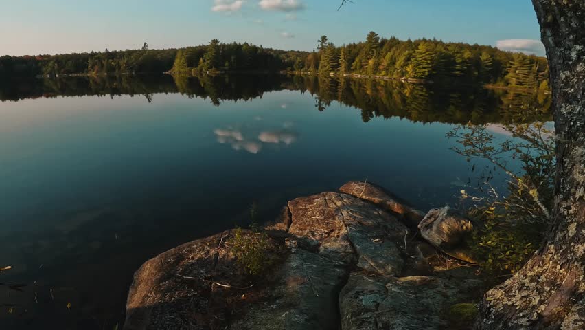 Golden-hour light reflecting off a still lake in Algonquin Park, Ontario, as clouds, trees, and rocky shoreline blend into a peaceful wilderness view