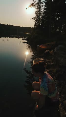 Young woman sits by calm lakeshore at sunset in Algonquin Park, Ontario with canoe in background
