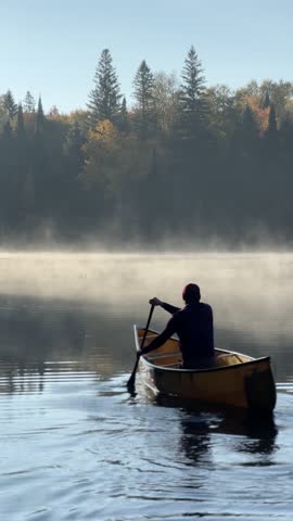 A Hispanic man paddles solo canoe across misty lake in Algonquin Park, Ontario at calm morning light