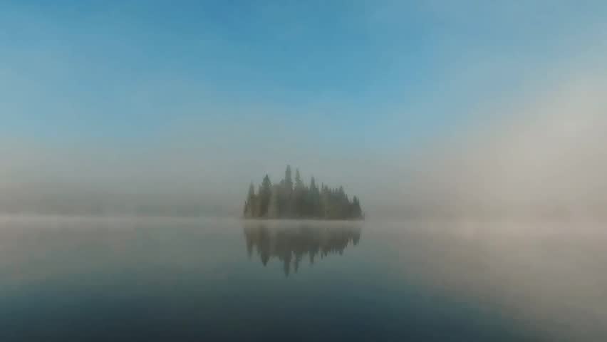 A Misty island with evergreen trees reflects in a calm lake under a soft, clear sky, shrouded in ethereal fog in Ontario, Algonquin Park, Canada