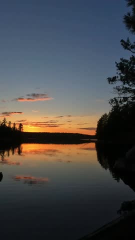 A calm lake at dusk reflects a vibrant orange and yellow sunset. The silhouettes of trees line the water