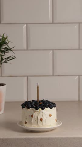 small bento cake decorated with fresh blueberries and almond slices on white plate with single golden candle burning against tiled kitchen wall, concept of birthday celebration, party, confectionery