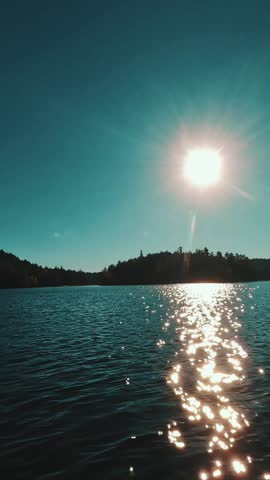 A vertical view of a backcountry forested shoreline and the sun shimmering across a glassy lake in Algonquin Park, Ontario, Canada