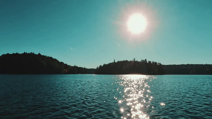 A view of a backcountry forested shoreline and the sun shimmering across a glassy lake in Algonquin Park, Ontario, Canada