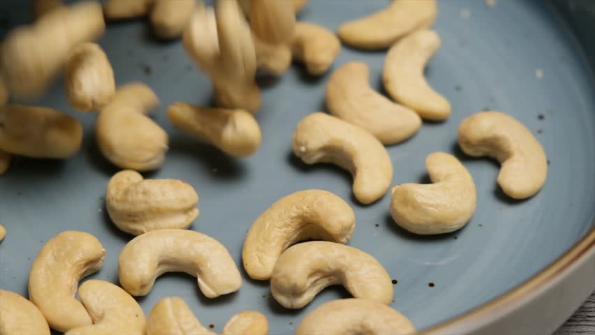 Close-up of golden cashews falling into a ceramic plate in slow motion. Ideal for healthy snacking, cooking, and adding crunch to salads, desserts, or trail mixes.