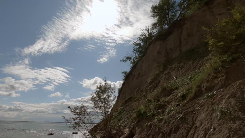 Slow-motion camera movement captures a natural, untouched shoreline at the base of a cliff. Gentle waves wash over rocks scattered along the sandy beach, creating a calm and cinematic coastal scene.