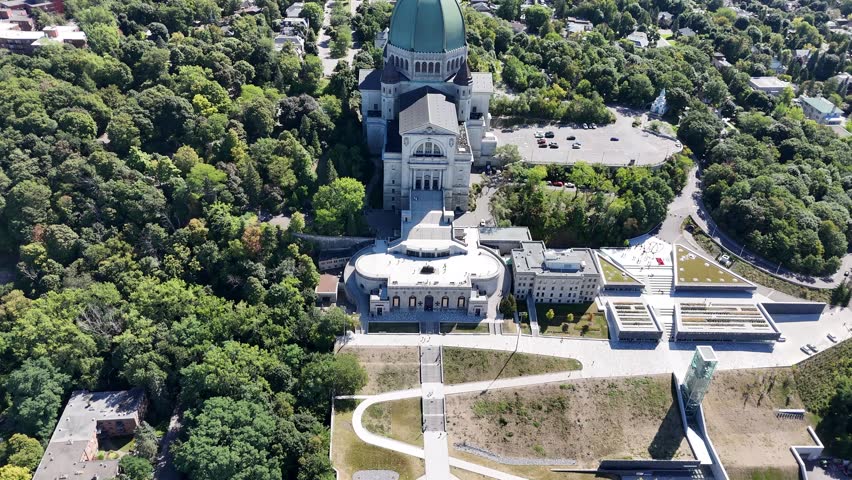 Drone flies forward from terrace and parking to dome, passes cross, pans up revealing Montreal skyline, bridges, and the St. Lawrence River