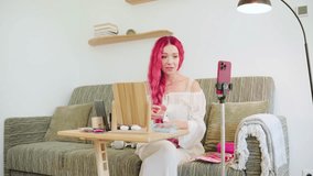 A woman with vibrant pink hair is applying makeup in her living room. She is filming a tutorial using a smartphone set on a tripod. The space is bright and inviting. - Powered by Shutterstock - Get 15% off with code: PIKWIZARD15