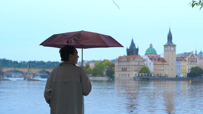 Woman with umbrella contemplating charles bridge in prague. Tourist holding umbrella standing on riverbank, gazing at iconic charles bridge stretching river during overcast autumn day in prague