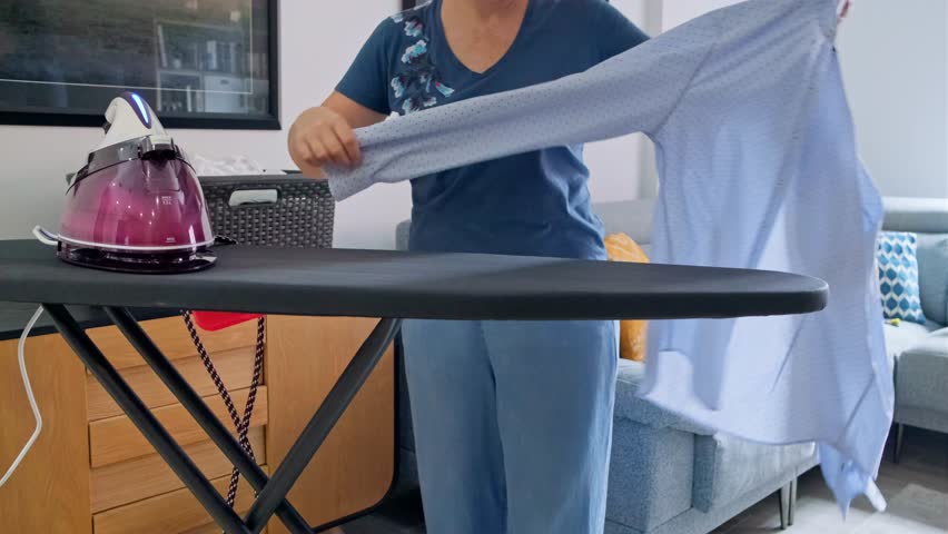 Unrecognizable housewife is ironing blue shirt on ironing board in living room, using steam iron, with a laundry basket and sofa in background. Common household chores concept.