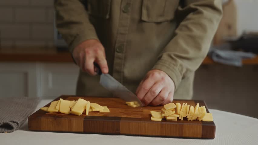 Man cutting block of yellow cheese into small pieces on wooden cutting board. Scene about balanced diet, dairy products, and homemade cooking. High quality 4k footage