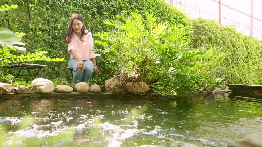 Happy asian woman sits relaxing and enjoying the peaceful atmosphere beside koi pond enjoying the lush nature of her home : Healthy wellness woman uses natural therapy maintain good mental health.