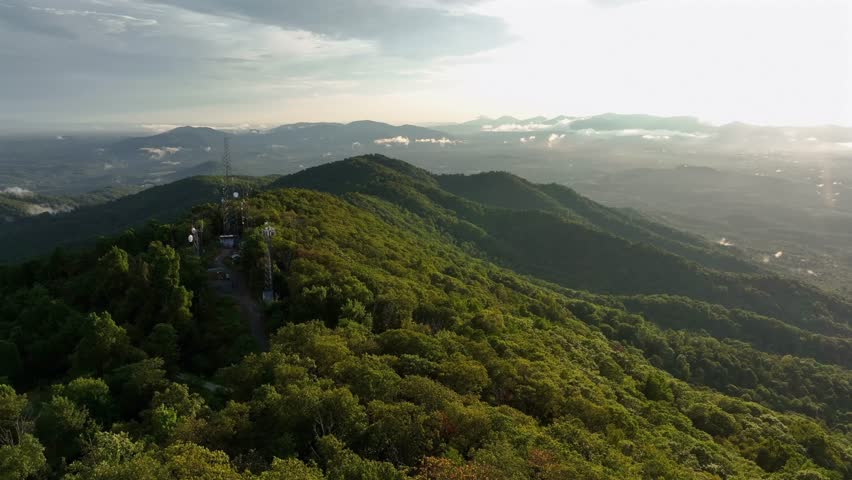 Radio tower or cell towers on peak of greened mountain ranges in USA. Peaceful sunrise over the clouds. Aerial wide shot. Panorama view in America.