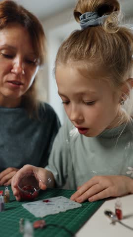 Goung girl concentrates while pouring red resin with glitter into silicone mold during home craft session. She explores her creativity under guidance of an adult.