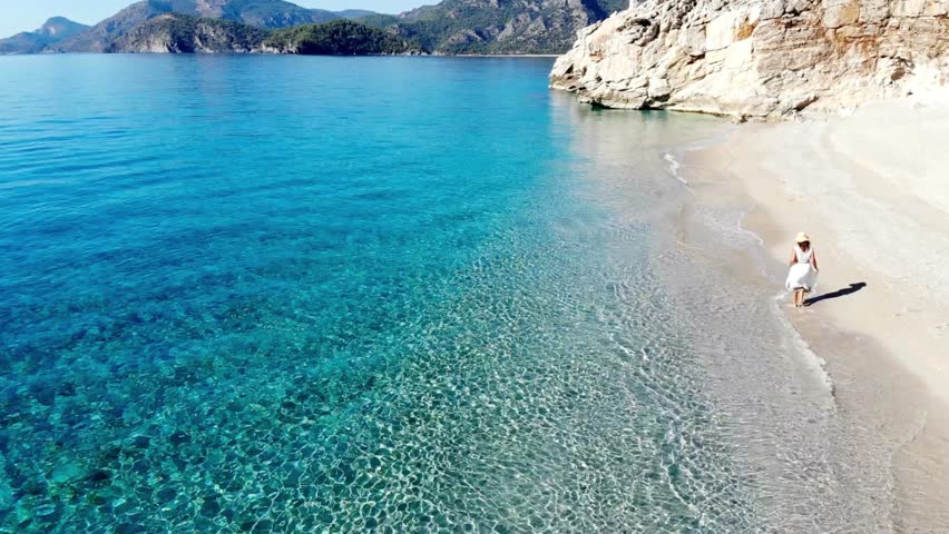 A woman in white dress walks along a pristine beach with turquoise water