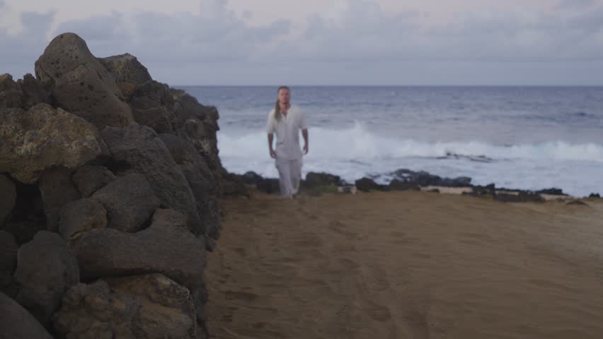 A man in loose white clothing walks confidently on a sandy coastal path beside a rugged stone wall, with the ocean horizon and waves stretching behind him.