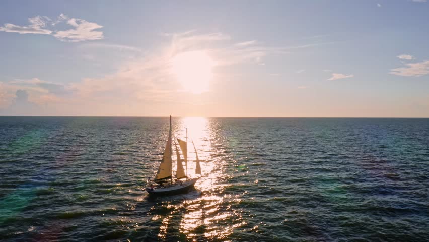 A sailboat glides smoothly over ocean waters beneath the afternoon sun, its sails casting shadows as golden reflections shimmer across the open sea.