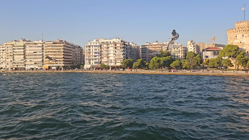 Thessaloniki seafront with the White Tower, residential buildings, trees, and people walking along the waterfront promenade beside the Aegean Sea under a clear blue sky