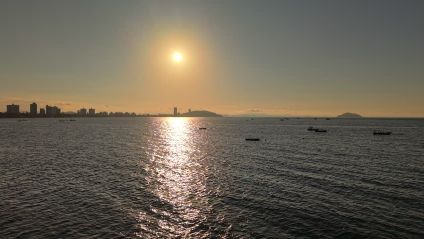A stunning aerial shot of the sunset over the ocean in Penha, Brazil. The golden sun reflects on the calm water, with a city skyline and small boats in the background.