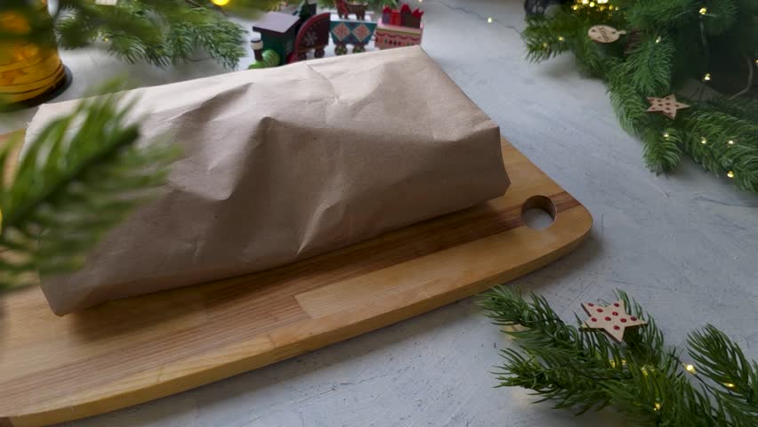 Static side view of female hands tying a gold ribbon around a stollen or gift wrapped in kraft paper on a festive background with Christmas elements, cozy seasonal scene.