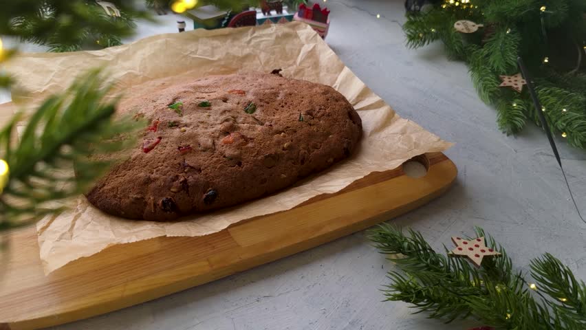 Static side view of female hands brushing melted butter over freshly baked stollen or fruit cake on parchment paper, with Christmas decor in the background.