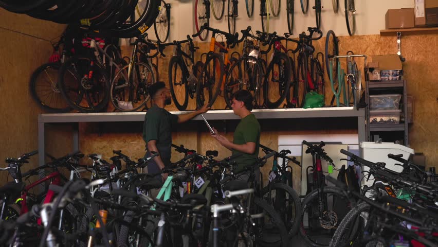 Professional bicycle mechanics carefully managing inventory in compact bike shop, collaborating while checking stock levels and pointing at organized bicycle parts and equipment