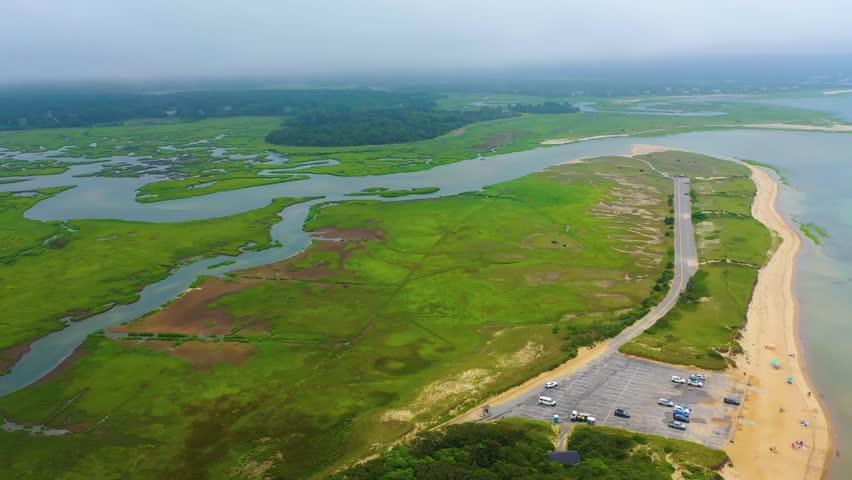 Drone flyover of marshland and coastline on a cloudy day, showing sandy beach, waterfront homes, winding tidal creeks, coastal greenery, and calm ocean waters stretching across the horizon.