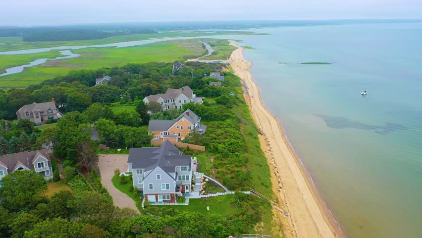 High-angle aerial shot of waterfront houses and expansive marshland beneath cloudy skies, with sandy beaches, winding tidal waterways, lush greenery, and calm waves rolling gently along the coast.