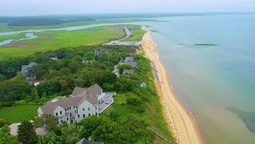 Scenic aerial view of oceanfront houses under cloudy summer skies, overlooking calm blue waters, sandy shoreline, tidal creeks, and expansive marshland stretching inland behind the coastal community.