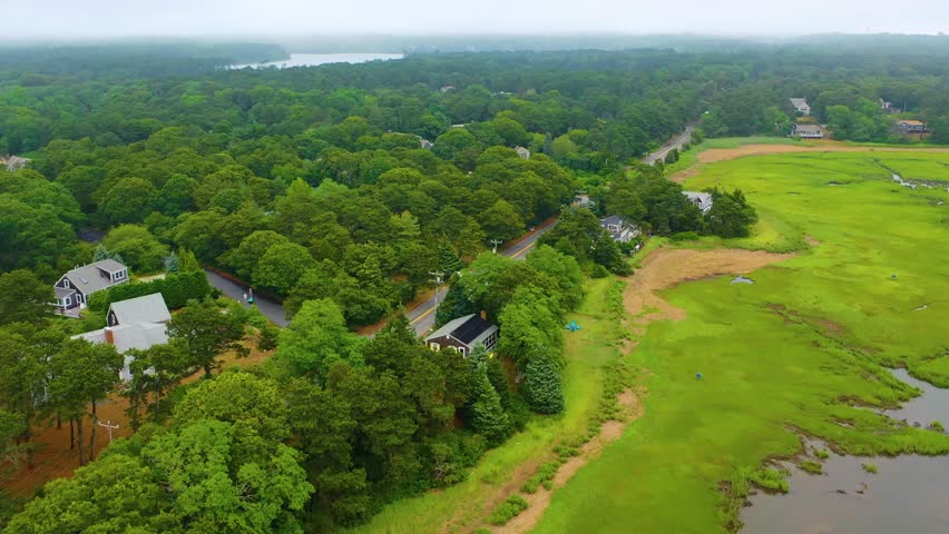 Aerial drone footage of Cape Cod marshlands, with winding tidal creeks flowing through lush green wetlands under an overcast summer sky, bordered by scattered homes and dense forest edges.