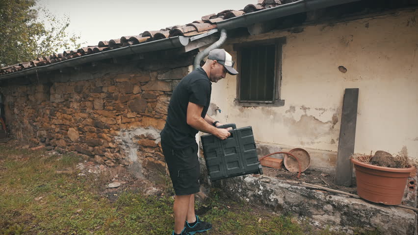A man unfolds and prepares a small hand truck cart next to the wall of a rustic countryside house. He adjusts the handles and sets the cart into position for use.