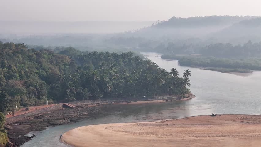 Misty morning aerial view of a wide river winding through a dense tropical jungle valley in the Konkan region of India.
