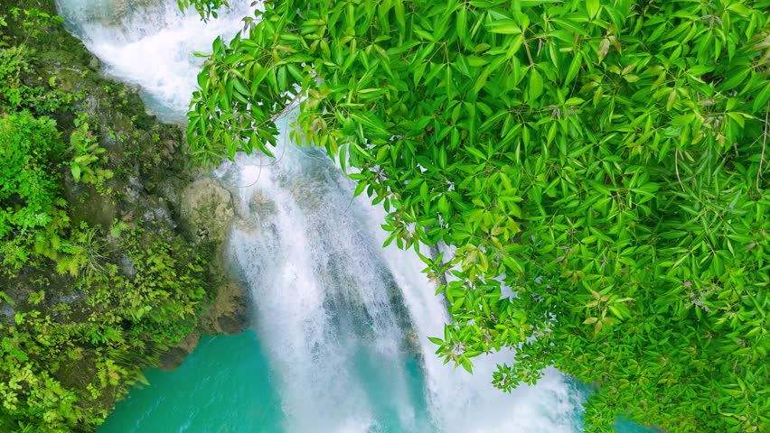 Aerial view of a multi-tiered waterfall cascading into turquoise pools, surrounded by jungle, with people swimming at Kawasan Falls, Philippines.