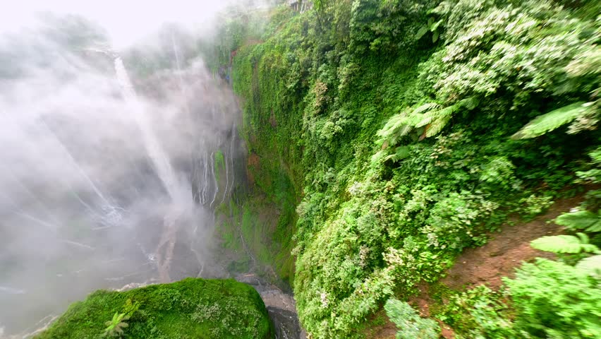 Cinematic FPV drone shot diving through the stunning multi-stream Tumpak Sewu waterfall, surrounded by lush rainforest cliffs in East Java, Indonesia.