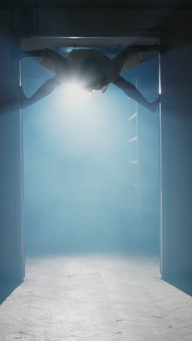 Terrifying silhouette of contortionist girl in hospital gown crawling and jumping in dark corridor with fog and cold backlight creating creepy Halloween horror scene in slow motion