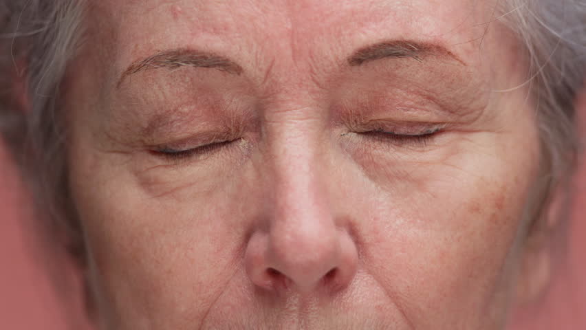 Detailed macro shot of an elderly woman face with eyes closed, showing wrinkles, skin texture, and natural aging features, isolated on pink background.