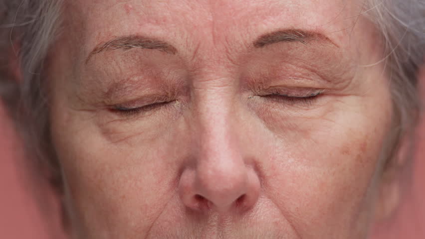 Detailed macro shot of an elderly woman face with eyes closed, showing wrinkles, skin texture, and natural aging features, isolated on pink background.
