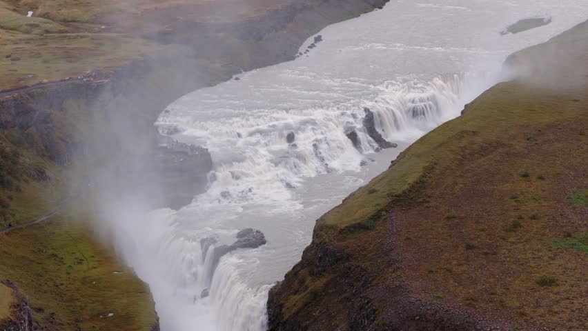 Drone captures Gullfoss waterfall in Iceland, with water cascading in two tiers into a canyon, mist rising, and grassy plains surrounding the scene.