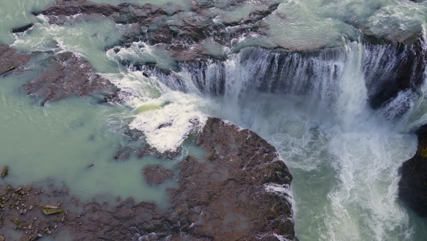 Aerial view of a multi tiered waterfall flowing into a turquoise river, surrounded by volcanic terrain, mossy greenery, and sparse vegetation.