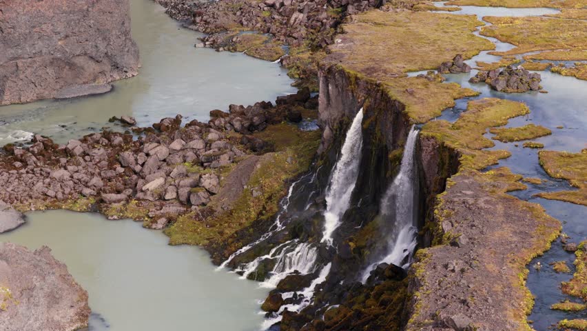 Aerial view of double waterfalls flowing over a rocky cliff into a river, surrounded by mossy volcanic terrain with winding streams in Iceland.