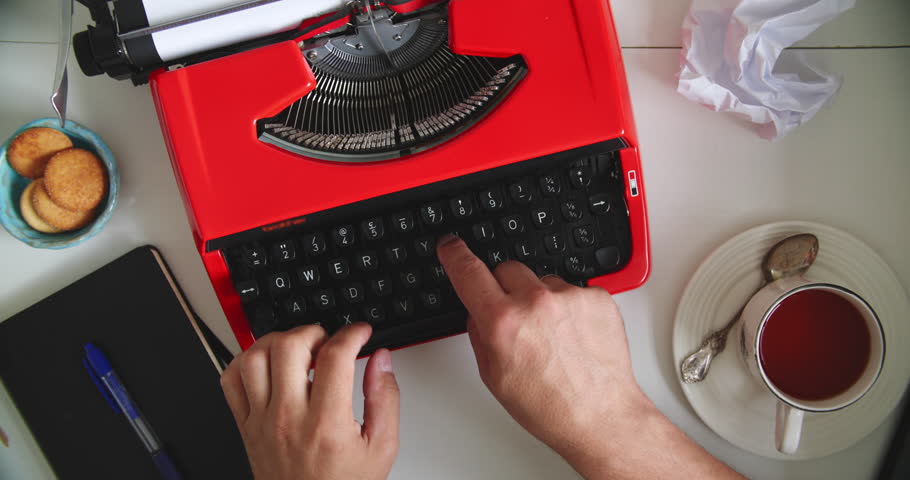 Hands typing on vintage red typewriter at desk with tea, cookies, notebook and crumpled paper. Concept of writing, creativity and retro style.