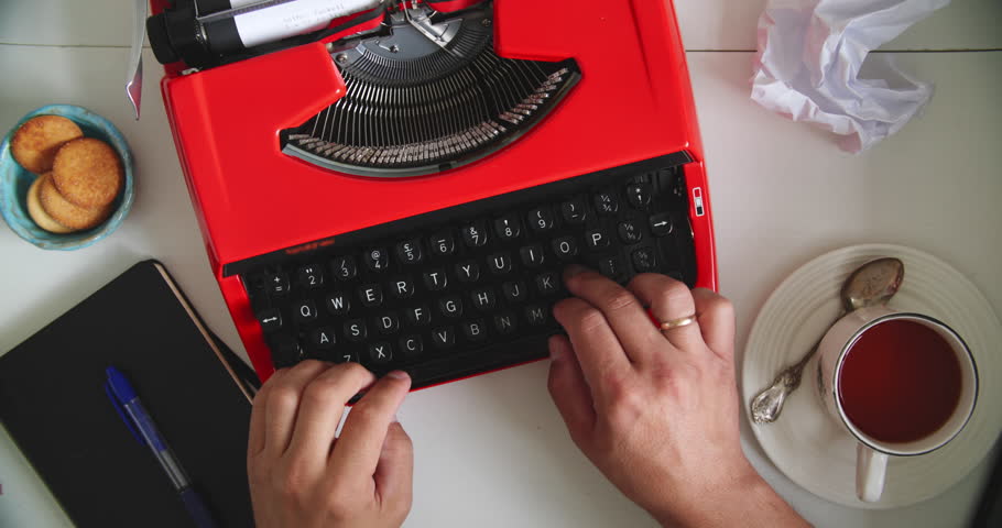 Hands typing on vintage red typewriter at desk with tea, cookies, notebook and crumpled paper. Concept of writing, creativity and retro style.