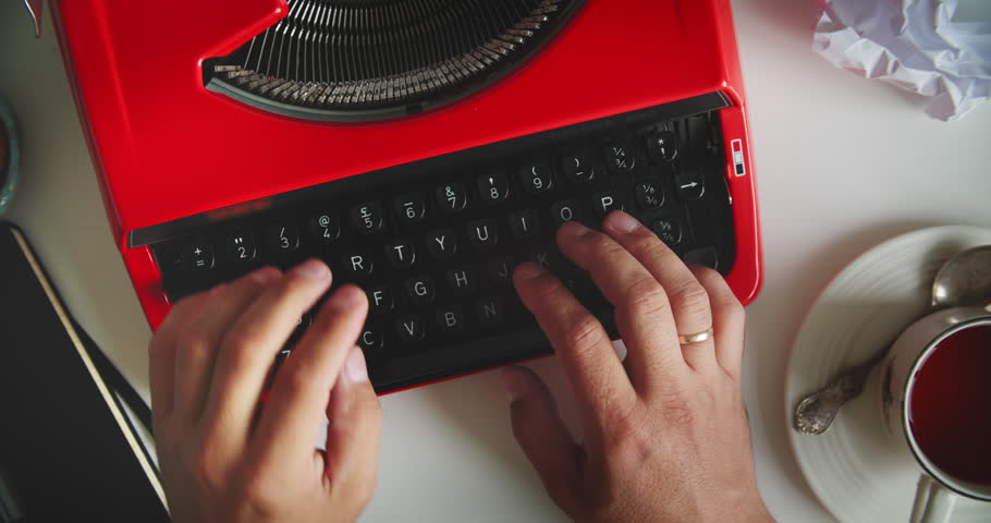 Hands typing on vintage red typewriter at desk with tea, cookies, notebook and crumpled paper. Concept of writing, creativity and retro style.