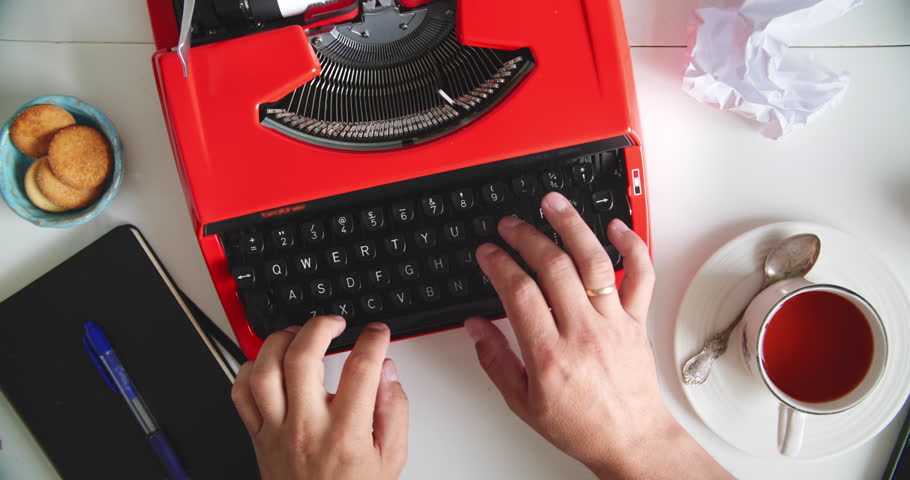 Hands typing on vintage red typewriter at desk with tea, cookies, notebook and crumpled paper. Concept of writing, creativity and retro style.