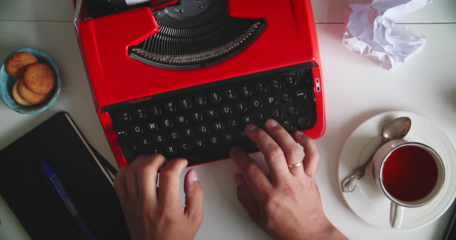 Hands typing on vintage red typewriter at desk with tea, cookies, notebook and crumpled paper. Concept of writing, creativity and retro style.
