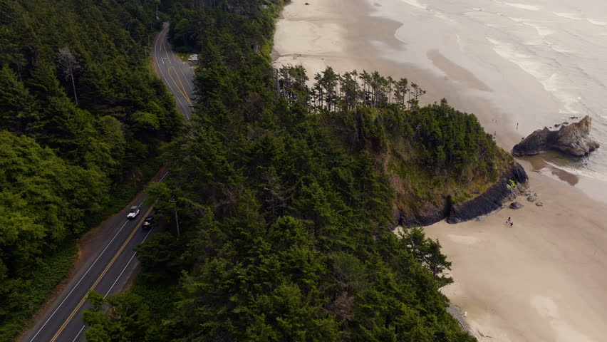 Drone aerial over Oregon Coast Highway 101 along Arcadia Beach, following cars driving past Douglas fir trees and shoreline with ocean waves on a clear day.