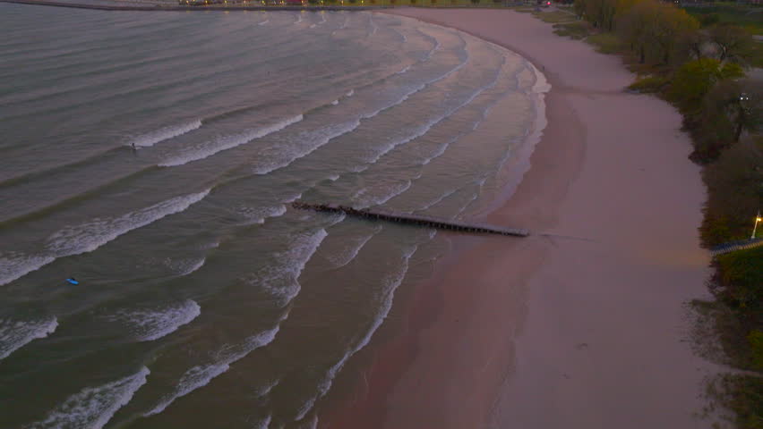 Drone aerial lowering over Lake Michigan shoreline in Sheboygan, Wisconsin at dusk with waves crashing on shore as surfers ride in the evening light.