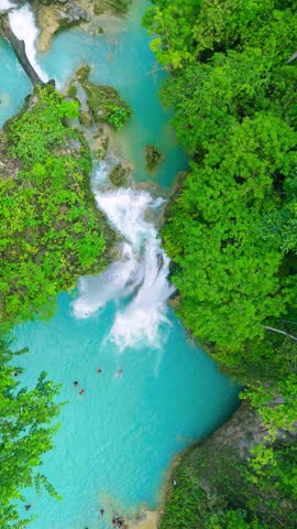 Aerial view of people swimming in the turquoise waters below a cascading waterfall, surrounded by lush green forest, Kawasan Falls, Philippines.