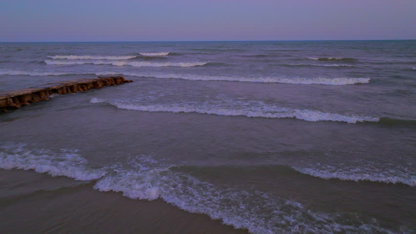 Drone aerial pulling away from gentle waves rolling onto the Lake Michigan shoreline in Wisconsin at dusk with soft evening light on the horizon.