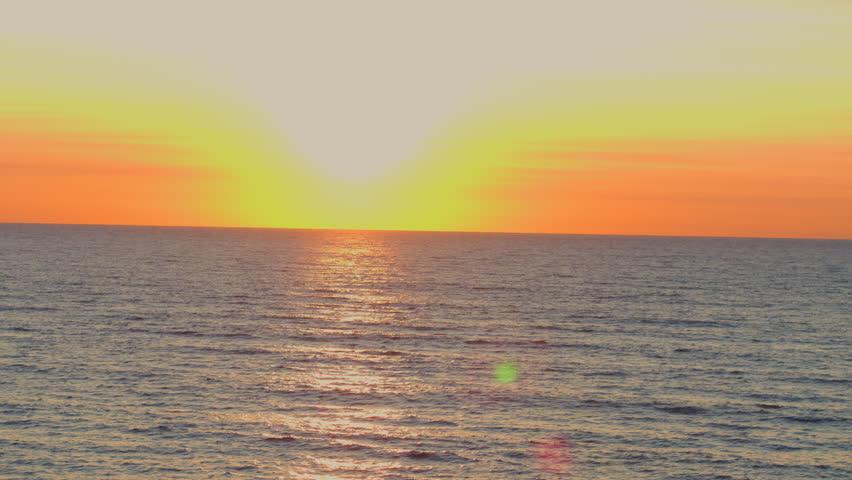 Drone aerial with a slow rise over Lake Michigan at sunrise, revealing gentle waves and a glowing horizon under a colorful morning sky.